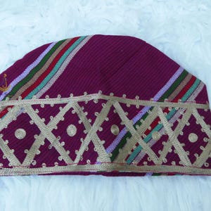 May include: A burgundy cap with a geometric gold design along the bottom edge. The cap features diagonal stripes in red, green, and purple. A small gold emblem is visible on the side. The cap is likely a cultural or religious head covering.