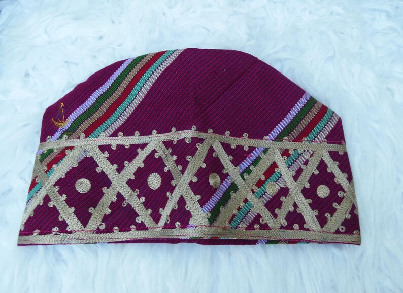May include: A burgundy cap with a geometric gold design along the bottom edge. The cap features diagonal stripes in red, green, and purple. A small gold emblem is visible on the side. The cap is likely a cultural or religious head covering.