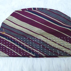 May include: A maroon and gold striped beanie hat with a small gold emblem. The hat features a pattern of diagonal stripes in maroon and gold, with a section of white polka dots. The hat is made of a soft, textured fabric.