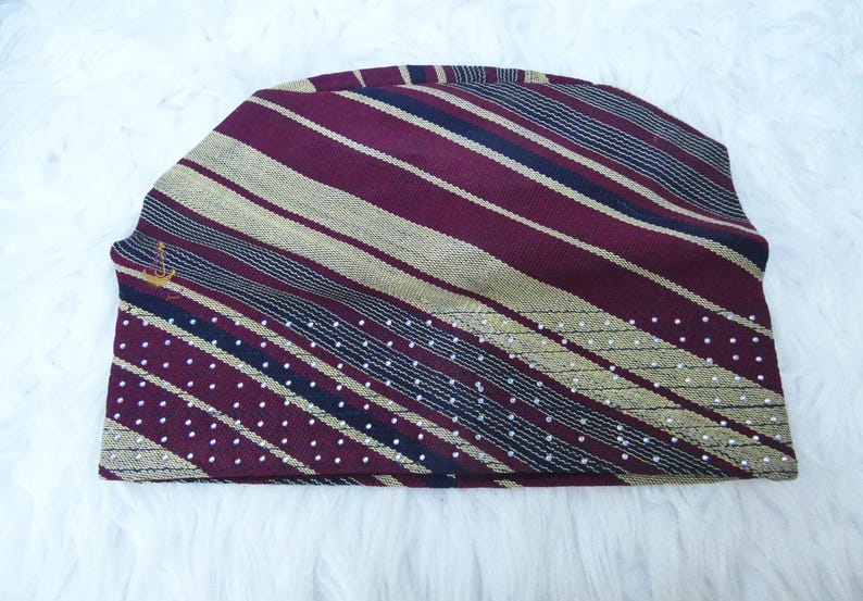 May include: A maroon and gold striped beanie hat with a small gold emblem. The hat features a pattern of diagonal stripes in maroon and gold, with a section of white polka dots. The hat is made of a soft, textured fabric.