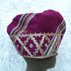 May include: A magenta-colored hat with a decorative gold trim and a geometric pattern. The hat features stripes of red, green, and white. A small gold emblem is visible on the side.