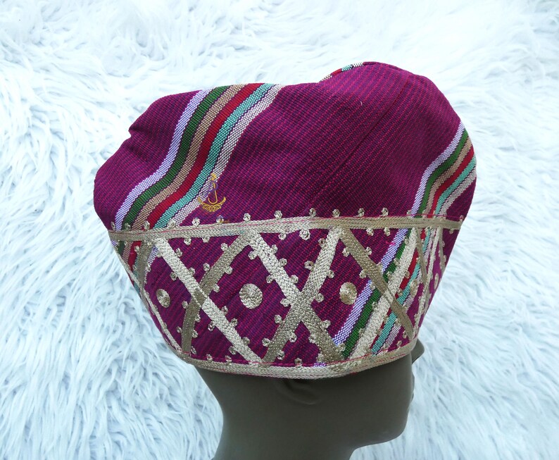 May include: A magenta-colored hat with a decorative gold trim and a geometric pattern. The hat features stripes of red, green, and white. A small gold emblem is visible on the side.