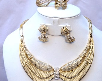 May include: Gold-tone jewelry set with a necklace, earrings, and a bracelet. The pieces are all made of a delicate, openwork design with a bow shape. The jewelry is accented with clear rhinestones.