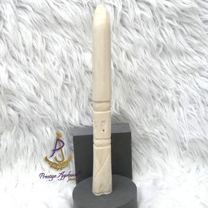 Puede incluir: Un objeto tallado de color marfil con una parte superior redondeada y un diseño texturizado detallado. El objeto está de pie sobre una base circular gris oscuro, con un bloque rectangular gris oscuro detrás. El objeto tiene un patrón geométrico tallado.