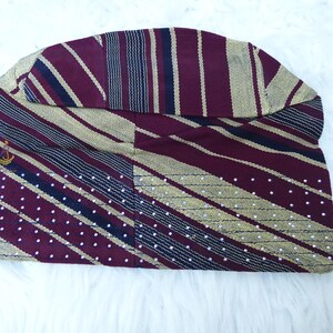 May include: A maroon and gold patterned hat with a small gold emblem. The hat features stripes, polka dots, and a curved brim. The fabric has a textured appearance, and the hat is displayed on a white surface.