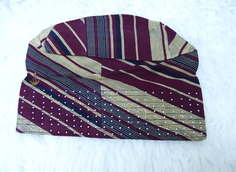 May include: A maroon and gold patterned hat with a small gold emblem. The hat features stripes, polka dots, and a curved brim. The fabric has a textured appearance, and the hat is displayed on a white surface.