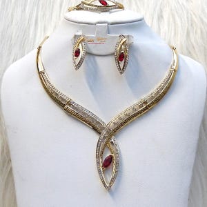 Puede incluir: Un conjunto de joyas dorado que incluye collar, pendientes y pulsera. Cada pieza tiene una gema roja central y está adornada con cristales transparentes. El collar y la pulsera presentan un diseño geométrico.