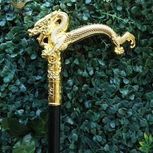 May include: A walking stick featuring a metallic gold-colored handle shaped like a dragon with intricate scale detailing. The handle sits atop a smooth, straight black shaft. The dragon head faces forward with a curved body forming the grip.