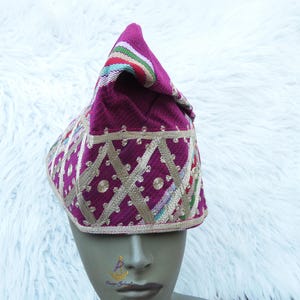 May include: A magenta-colored, traditional hat with a pointed crown. The hat features gold-colored geometric embroidery and a striped band of color at the top. The hat is displayed on a mannequin head.