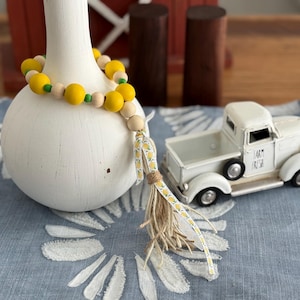 May include: A white ceramic vase is adorned with a yellow and natural wood bead garland. A small white toy truck with the words "Farm Fresh" is on a blue floral patterned cloth. The garland has a tassel with a yellow lemon ribbon.