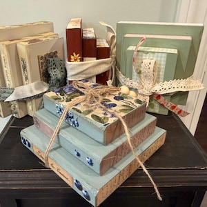 May include: A collection of decorative wooden blocks designed to resemble stacked books. The set includes various stacks tied with twine, lace, and fabric ribbons, featuring painted finishes in shades of green, cream, and brown with floral and script patterns.