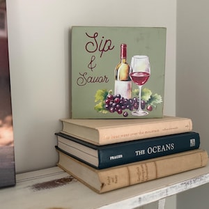 May include: A decorative square sign with a wine theme, featuring a bottle, a glass of red wine, and grapes. The sign is stacked on top of three books. The sign reads "Sip & Savor" in cursive script.