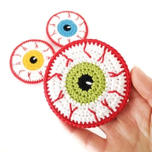 May include: Three crocheted eye-shaped coasters. The coaster in the foreground has a green iris, a black pupil, and red veins. The other two coasters have blue and yellow irises.