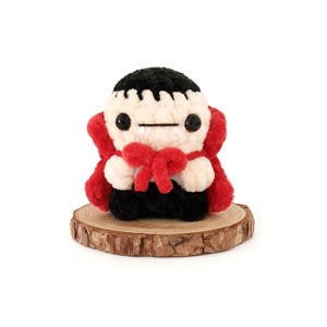 May include: A small, crocheted vampire plush toy with a red cape and a black bow tie. The toy is sitting on a wooden base.