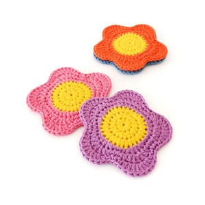 May include: Three handmade crochet flower coasters in bright colours. One is orange with a yellow centre and blue trim, another is pink with a yellow centre, and the third is purple with a yellow centre. These decorative coasters are perfect for adding a touch of colour to any room.
