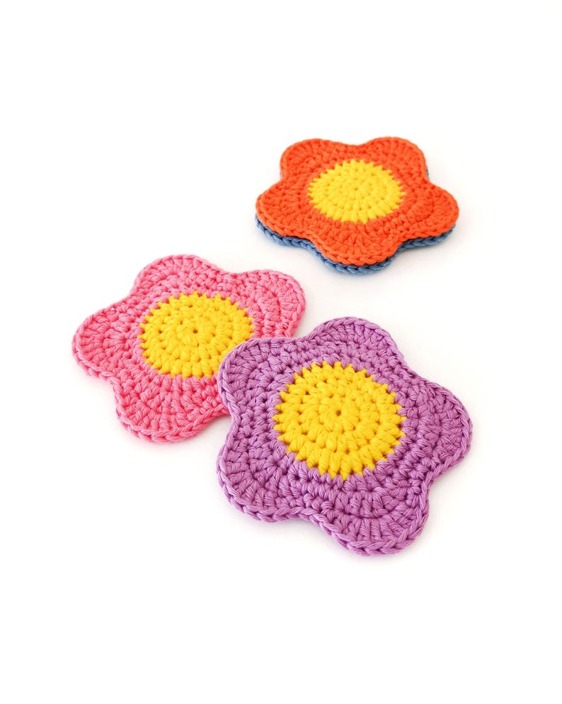 May include: Three handmade crochet flower coasters in bright colours. One is orange with a yellow centre and blue trim, another is pink with a yellow centre, and the third is purple with a yellow centre. These decorative coasters are perfect for adding a touch of colour to any room.