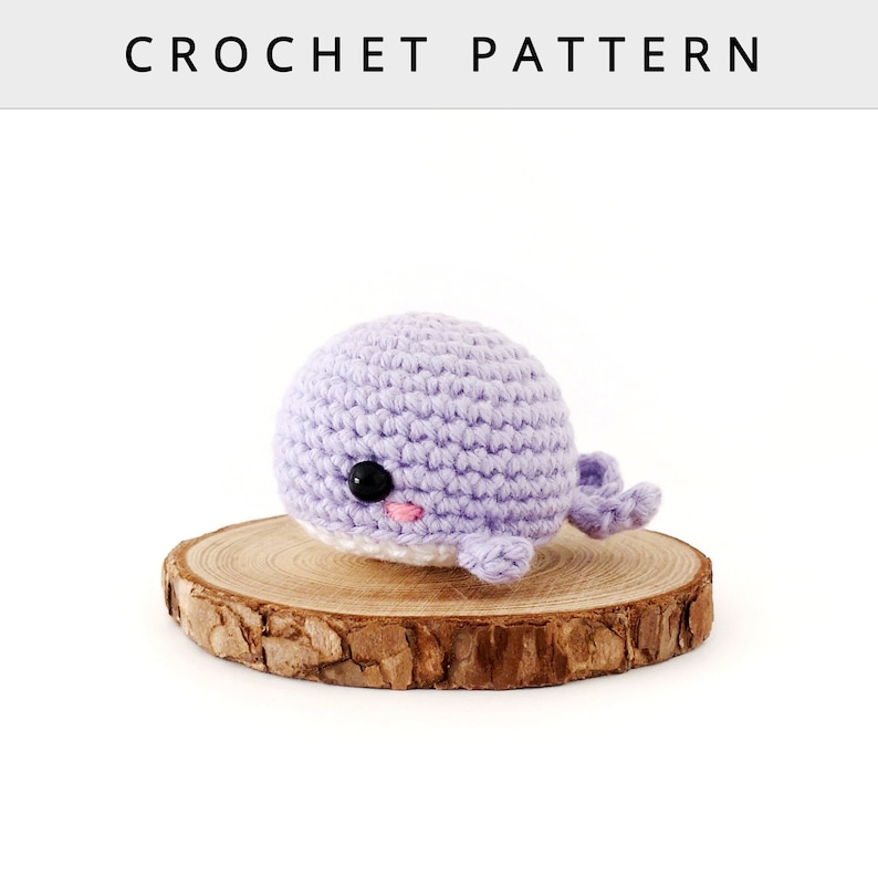 May include: A crocheted whale amigurumi toy with a purple body, white belly, and black eyes. The whale is sitting on a wooden slice.