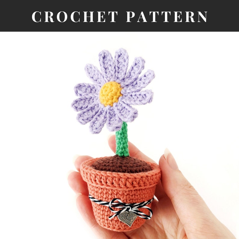 May include: A crocheted purple flower with a yellow center in a terracotta pot with a brown and white ribbon. The pot has a small silver charm with a heart on it.