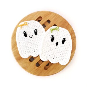 Puede incluir: Dos adornos de fantasmas de crochet blanco con ojos y bocas negros. Los fantasmas tienen lazos amarillos y verdes en la parte superior de sus cabezas. Los fantasmas est&aacute;n sentados sobre una superficie de madera.