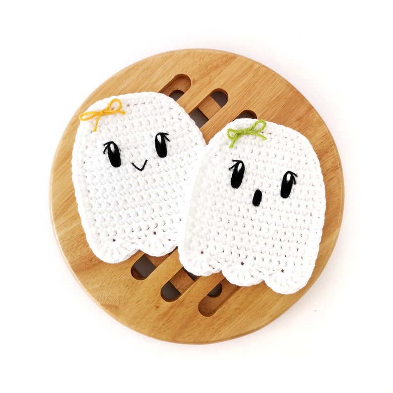 Puede incluir: Dos adornos de fantasmas de crochet blanco con ojos y bocas negros. Los fantasmas tienen lazos amarillos y verdes en la parte superior de sus cabezas. Los fantasmas est&aacute;n sentados sobre una superficie de madera.