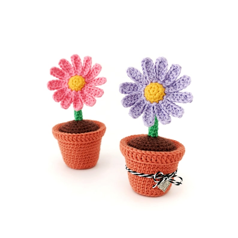 May include: Two crocheted flower pots with pink and purple flowers. The pots are orange and have brown soil. The purple flower pot has a black and white ribbon and a silver tag with the word "Love" on it.