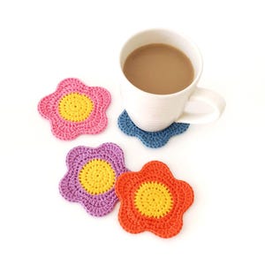 May include: A white mug containing a light brown beverage rests on a blue crocheted flower coaster. Three more crocheted flower coasters in pink, purple, and orange with yellow centres are also shown. The coasters are handmade.
