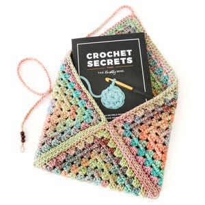 Op de afbeelding: Een gehaakte enveloptas in een granny square patroon, met een boek getiteld "Crochet Secrets from The Knotty Boss". De tas is gemaakt van pastelkleurig garen en heeft een roze band met houten kralen. Afmetingen: ongeveer 25 cm.