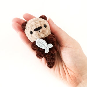 May include: A small crocheted otter plush toy holding a blue fish. The otter is brown and beige with black eyes and a black nose.