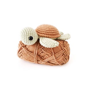 May include: A crocheted turtle in shades of tan, light green, and brown, resting on a ball of yarn. The turtle has a tan shell, light green flippers, and a light green head with a black button eye. The yarn is a warm, earthy tone.