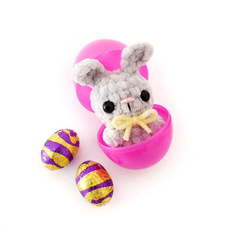 May include: A gray crocheted bunny rabbit sits inside a pink plastic Easter egg. Two foil-wrapped chocolate Easter eggs are on a white surface.