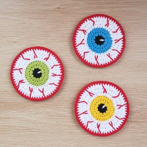 May include: Three crocheted eye-shaped coasters with red, white, and different coloured centres. The coasters are on a light brown wooden surface.