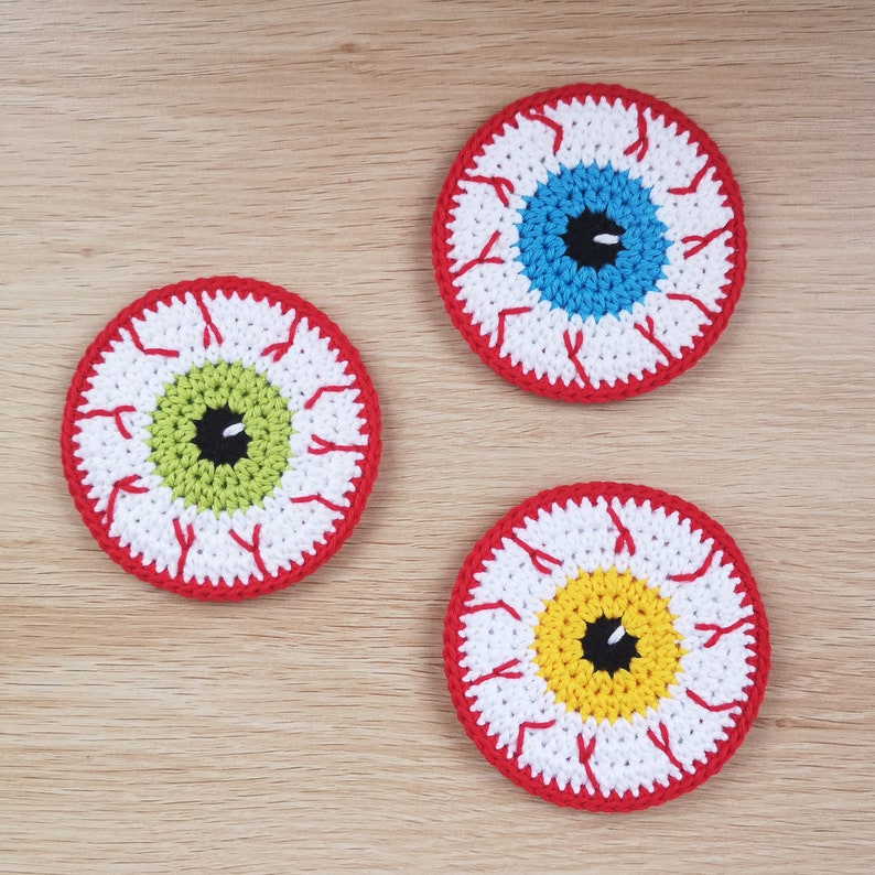 May include: Three crocheted eye-shaped coasters with red, white, and different coloured centres. The coasters are on a light brown wooden surface.