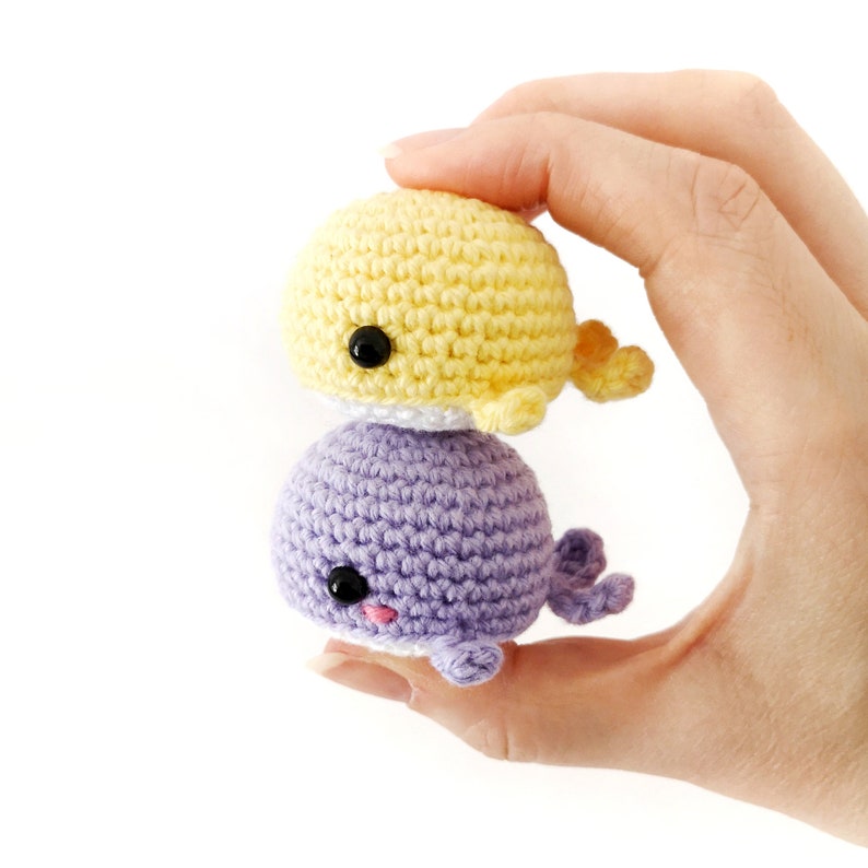 May include: Two crocheted whale plushies, one yellow and one purple, stacked on top of each other. The whales have black eyes and pink noses.