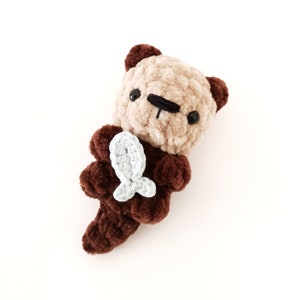 May include: A brown and beige crocheted stuffed otter holding a light blue crocheted fish.