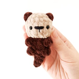 May include: A small crocheted stuffed otter with a brown body and a beige head. The otter has black eyes and a black nose.