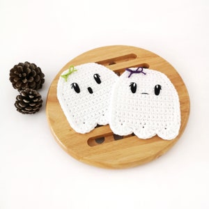 Puede incluir: Dos posavasos de fantasma de crochet blanco con ojos y bocas negros. Un fantasma tiene un lazo verde y el otro un lazo morado. Los posavasos est&aacute;n sobre una superficie de madera.