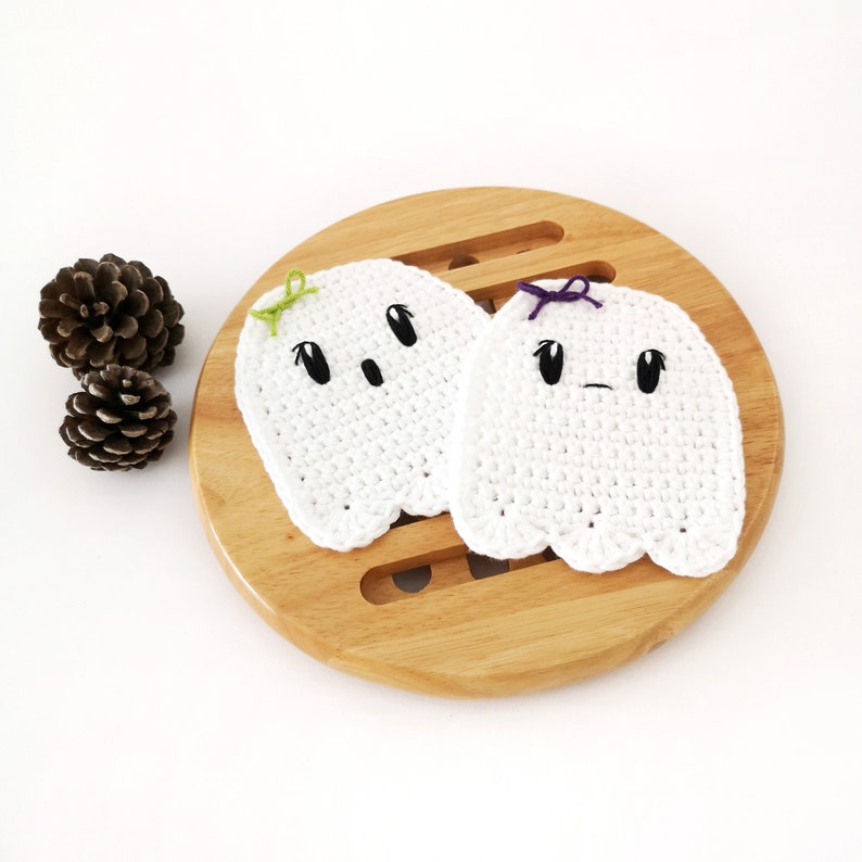 Puede incluir: Dos posavasos de fantasma de crochet blanco con ojos y bocas negros. Un fantasma tiene un lazo verde y el otro un lazo morado. Los posavasos est&aacute;n sobre una superficie de madera.