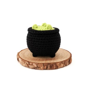 May include: A small black crocheted cauldron with green crocheted goo inside, sitting on a wooden slice.