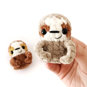 May include: Two crocheted sloth plushies, one brown and one gray with white accents. Both have black eyes and a smiling face.