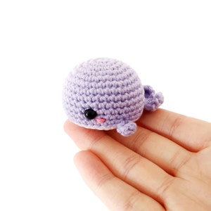 May include: A small, crocheted whale toy in a light purple color. The whale has a black eye and a pink mouth.