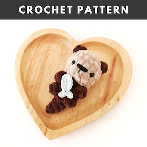 May include: A brown and beige crocheted otter holding a light blue crocheted fish. The otter is sitting on a wooden heart-shaped plate.