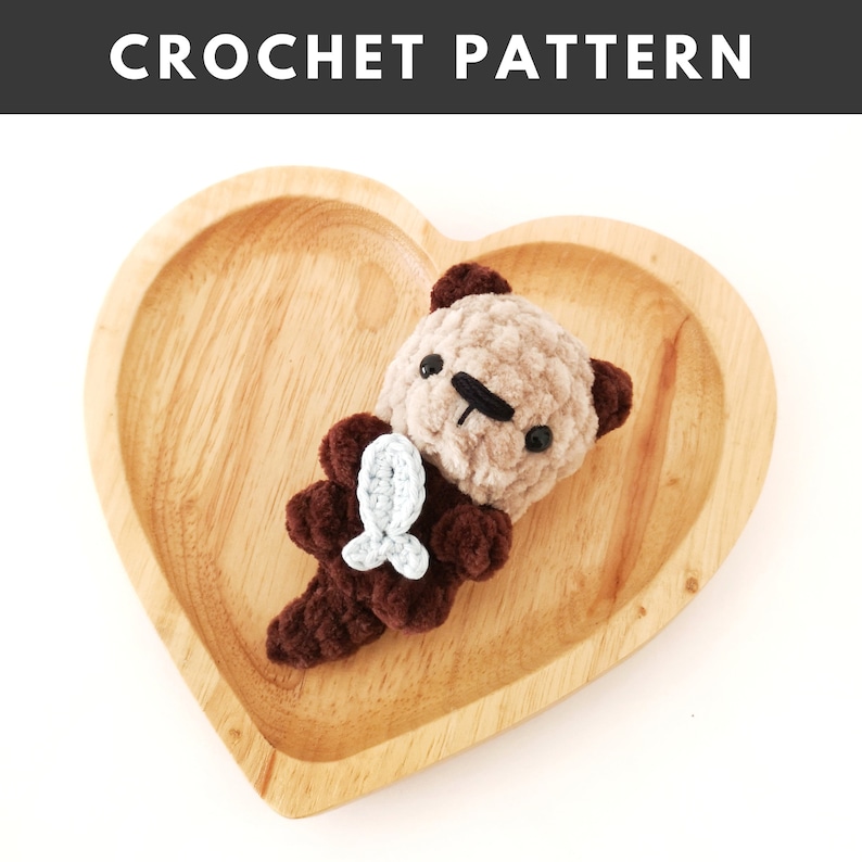 May include: A brown and beige crocheted otter holding a light blue crocheted fish. The otter is sitting on a wooden heart-shaped plate.