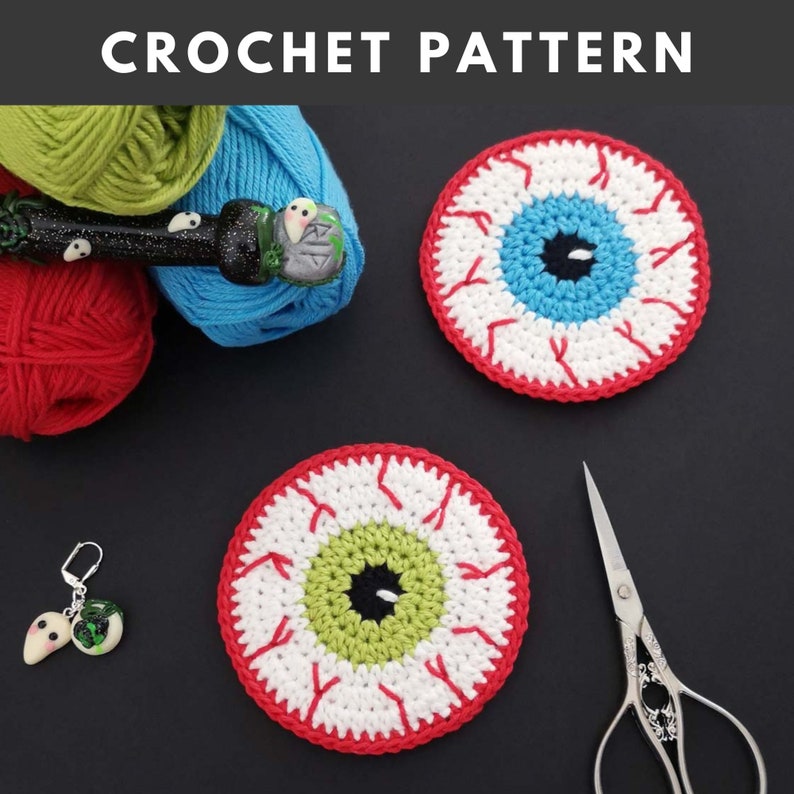 May include: Two crocheted eyeball coasters, one with a blue iris and one with a green iris. Both have red veins and white sclera. The coasters are on a black surface with yarn, a crochet hook, and scissors.