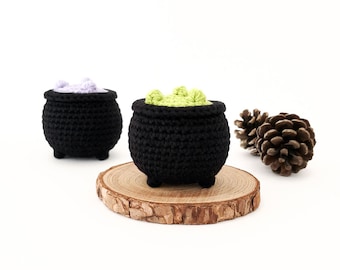 May include: Two black crocheted cauldrons with handles, one filled with green and the other with purple, sitting on a wooden slice. A pine cone is on the right side of the image.