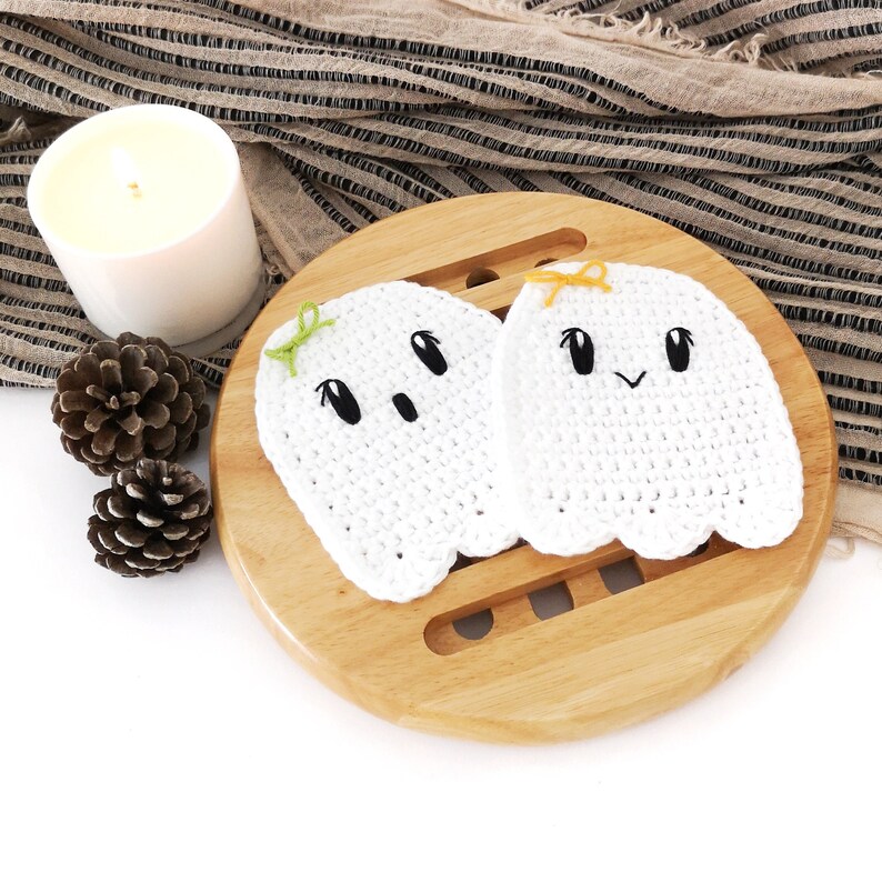 Puede incluir: Dos posavasos de fantasma de crochet blanco con ojos negros y una boca sonriente. Los posavasos est&aacute;n en una bandeja de madera.