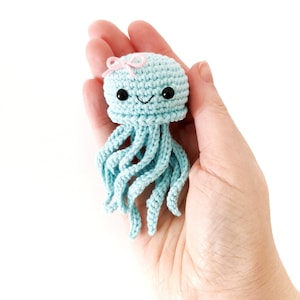 May include: A light blue crocheted jellyfish with a pink bow, black eyes, and a smiling mouth. The handmade sea creature has long, flowing tentacles. The jellyfish is held in a hand against a white background.