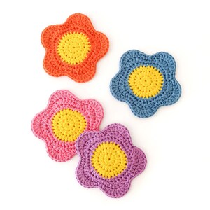 May include: Four colourful, crocheted flower-shaped coasters. The coasters are in shades of orange, blue, pink, and purple, each with a yellow centre. The handmade coasters are arranged on a white background.