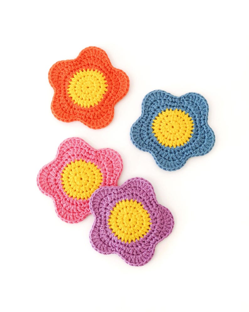 May include: Four colourful, crocheted flower-shaped coasters. The coasters are in shades of orange, blue, pink, and purple, each with a yellow centre. The handmade coasters are arranged on a white background.