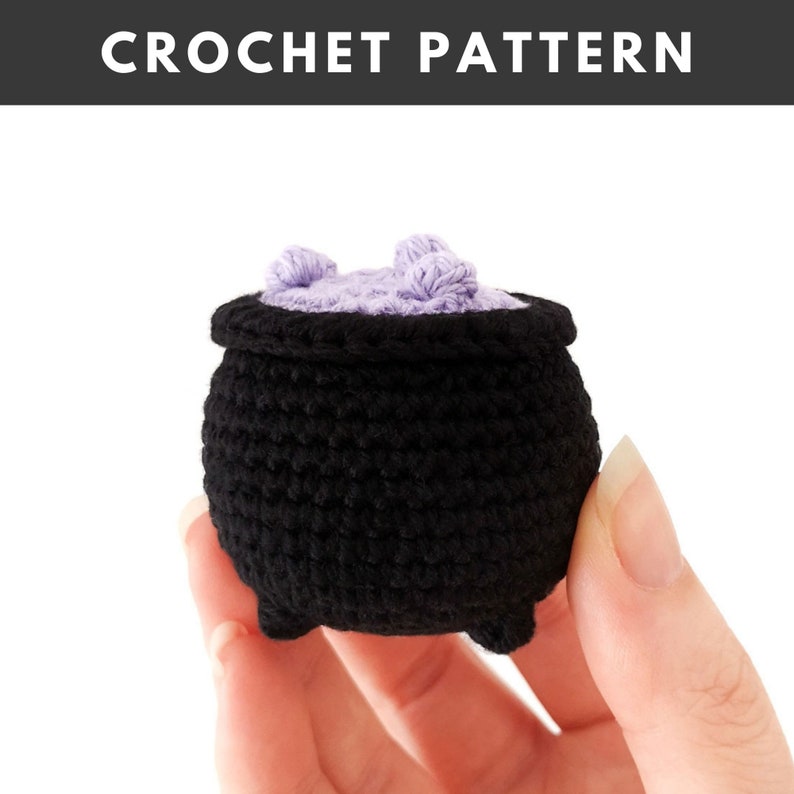 May include: A crocheted black cauldron with a purple lid and two purple pom-poms on top. The cauldron is being held in a hand.