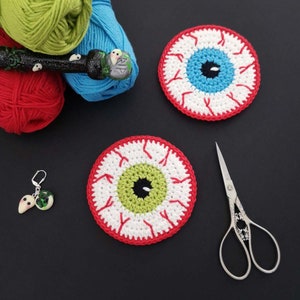 May include: Two crocheted eye-shaped coasters made with red, white and green yarn. The coasters are on a black surface with a pair of silver scissors.